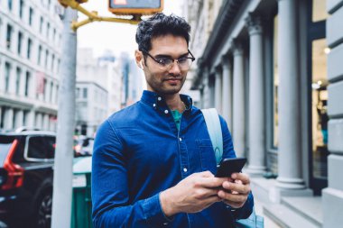 Gündüz vakti işlek caddede dikilirken akıllı telefondan mesaj atan, rahat tişörtlü ve gözlüklü kendinden emin, yakışıklı bir yetişkin adam.