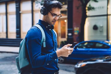 Kulaklıklı, sırt çantalı, akıllı telefon ekranlı gözlüklü, gündüzleri şehir caddesinde yürürken odaklanmış, yakışıklı, yetişkin bir adamın yan görüntüsü.