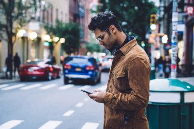 Kendine güvenen yakışıklı, ceketli ve gözlüklü yetişkin bir adamın akıllı telefon kullanarak kentsel yaya geçidinde durmasının yan görüntüsü.