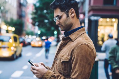 Sakin, yakışıklı, ceketli ve gözlüklü yetişkin bir adamın gündüz şeridinin yanında dururken akıllı telefon kullanmasının yan görüntüsü.
