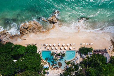 Açık yüzme havuzu ve güneş pansiyonları olan deniz manzaralı otel oteli. Yaz tatilinin kuş bakışı manzarası, turkuaz deniz suyu ve temiz kum kıyısındaki şemsiyeler