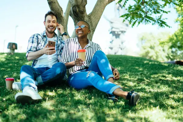 笑积极浪漫的多种族情侣在绿地绿地树荫下 一边与电话和咖啡一起玩乐 — 图库照片