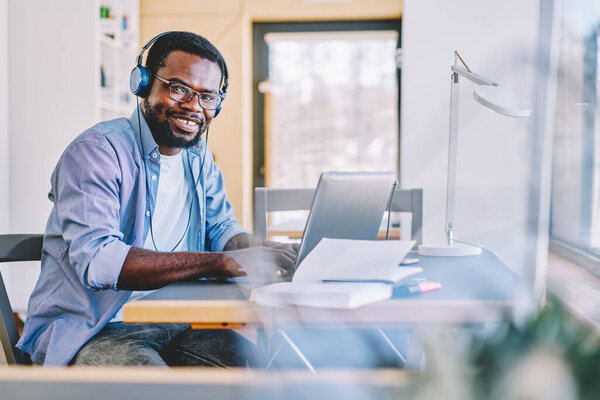 Cheerful African American man in headphones smiling and looking at camera while typing on laptop keyboard and listening to music in modern workplace