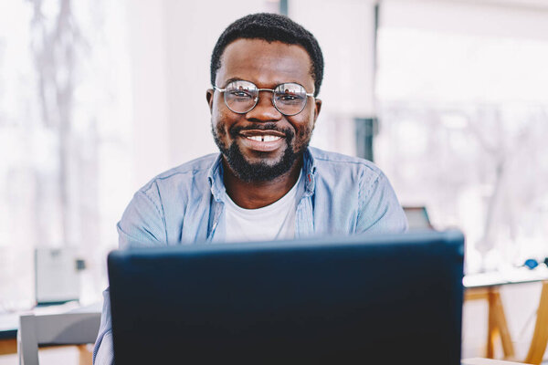 Cheerful smart adult bearded black man in glasses looking at camera and using laptop while sitting in light modern office