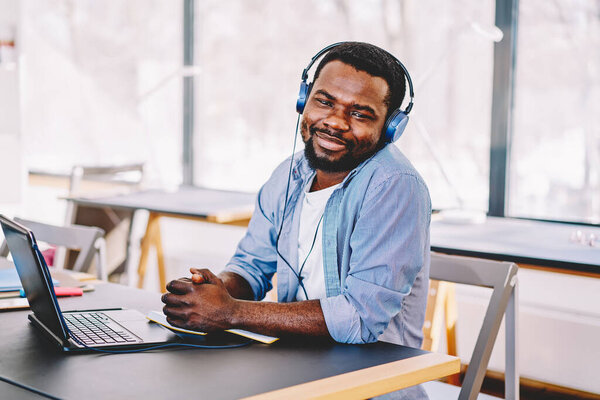 Satisfied successful African American male designer in headphones looking at camera while sitting with laptop at table in modern art studio