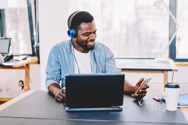 Happy successful black man in headphones using cellphone while working and sitting with laptop at table in light modern office