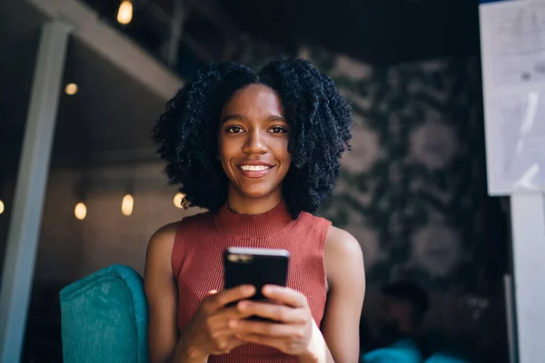 非洲裔美国人穿着休闲装 满脸笑容 卷曲的金发女士在现代咖啡馆里用智能手机查看社交媒体 — 图库照片