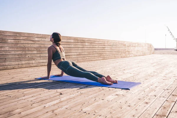 穿着运动服训练瑜伽的成年女子侧视图 在明亮的阳光下在木制长廊上做冲浪板 — 图库照片