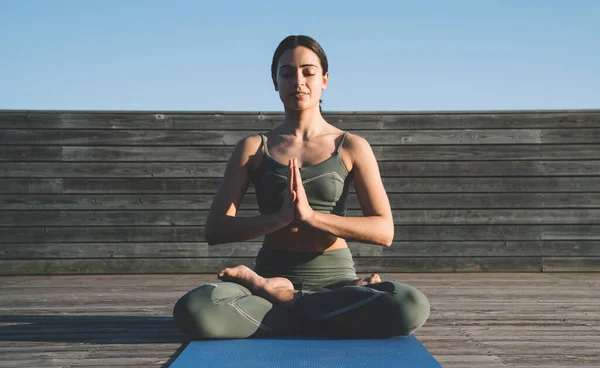 Slender serene ethnic woman sitting in lotus position peacefully with folded hands and closed eyes on mat on wooden pathway