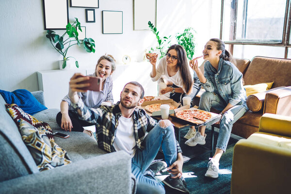 Cheerful group of friendly young people smiling and taking selfie on mobile phone while eating pizza at table sitting on sofa and floor with crossed legs 