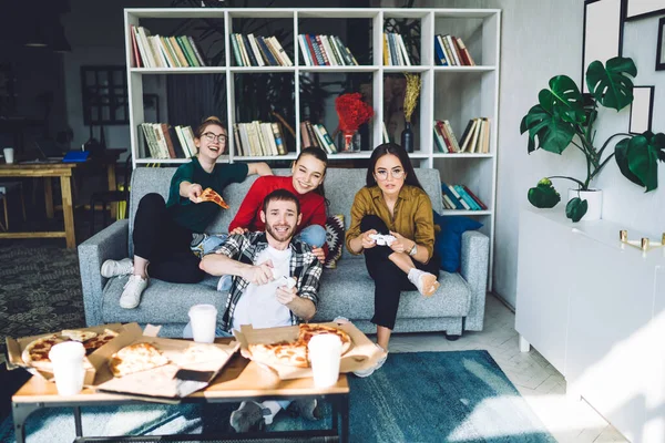 一群友善的年轻游戏玩家坐在有书架的客厅沙发上玩着游戏机和吃着披萨 — 图库照片