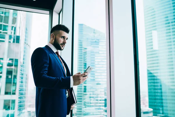 From below handsome serious executive man in elegant clothes looking away and making strategic plans while looking out window and interacting with smartphone in light contemporary office