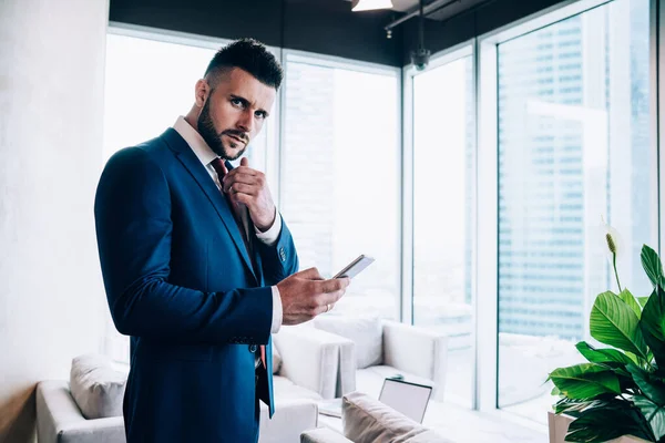 Concerned male coworker in blue formal suit rubbing chin in thoughts while surfing on mobile phone and looking at camera at office