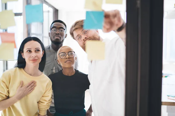Multiracial business people in casual outfit and glasses having conversation standing behind glass in office pasting sticky notes on glass wall sharing ideas. Collaboration in team, brainstorming