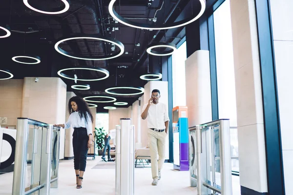 Confident smart African American partners in stylish wear passing through turnstile while communicating on smartphone in hall of modern business center