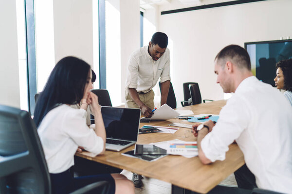 African American male executive in formal clothes standing in conference room and reading papers while explaining details of new project for employees 