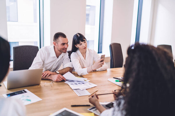 Glad and cheerful office employees smiling and having fun during business meeting while sitting in front of papers and browsing social media  