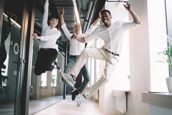 Diverse young excited enthusiastic fun business team jumping high with raised arms for celebration of successful work in modern office corridor