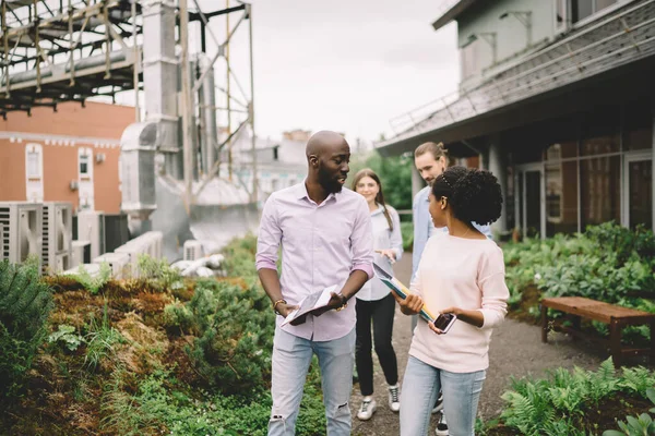 Group of multiethnic people walking on terrace of office and talking surrounded by bushy plants in background of city in daylight