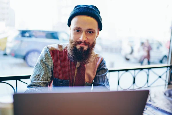 时尚的男人穿着五颜六色的衬衫 戴着黑色的温暖的帽子 白天坐在咖啡厅的阳台上 专注于屏幕 与笔记本电脑互动 — 图库照片