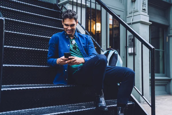 Cheerful modern man in stylish outfit sitting on street stairway and browsing mobile phone with smile on street of New York city