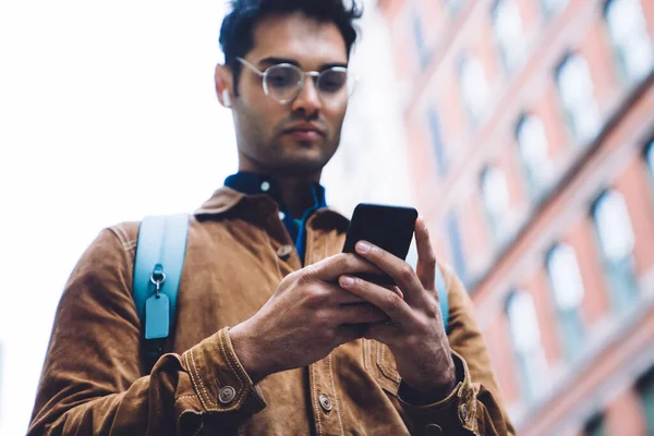 从下面的图片中可以看到 在纽约 身穿褐色羊皮夹克 背包和耳机 头戴眼镜 头戴眼镜 头戴墨镜的西班牙裔男性在出租汽车租赁大楼的背景下在手机上打字 — 图库照片