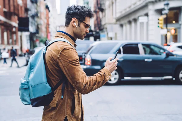 身穿褐色绒面革夹克 背着背包 带着耳机用手机发短信 一边在纽约市四处走走一边听音乐的西班牙裔年轻人的侧影 — 图库照片