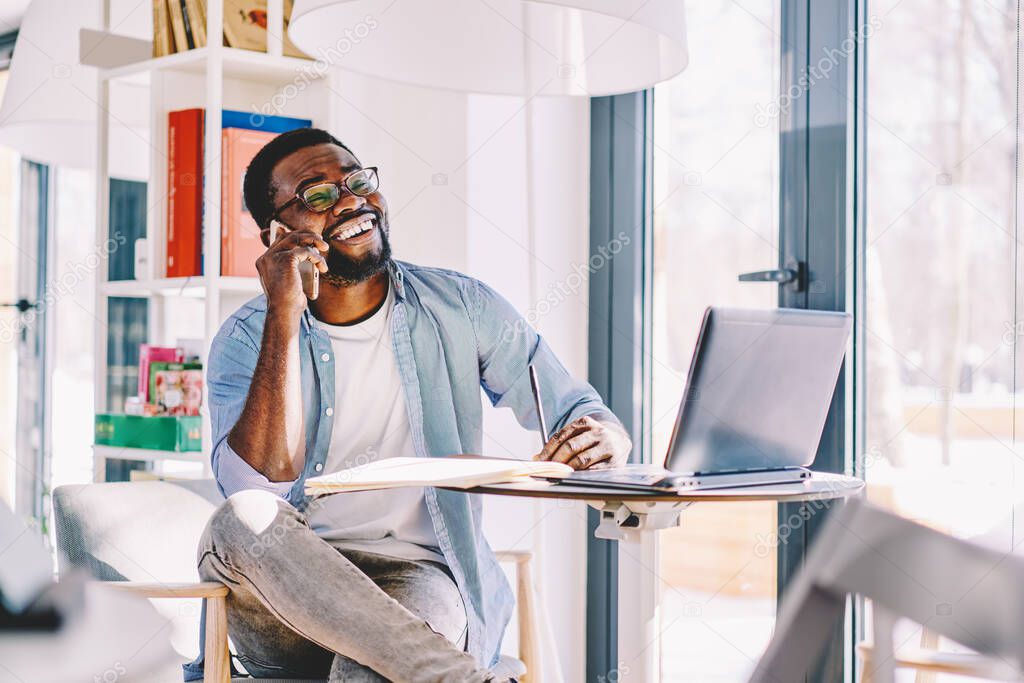 Pensive African American freelancer in glasses and casual clothes making phone call and laughing while sitting at table with papers and laptop at home
