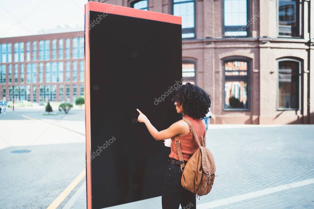Back view of black bright traveler with Afro hairstyle in casual wear with bag pointing at billboard walking in street