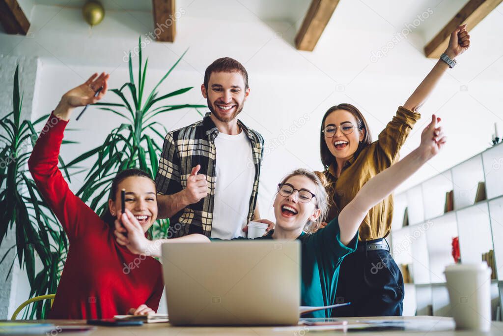Group of delighted classmates raising arms and smiling while looking at laptop after homework assignment fulfillment during studies at home