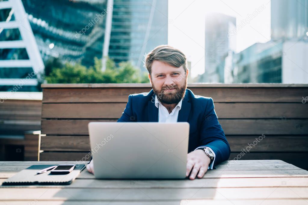 Handsome classy man in business suit working on laptop while sitting at table in street cafe on background of urban buildings