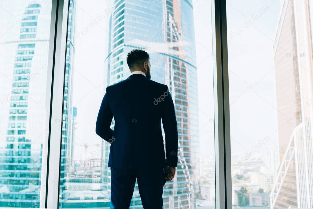 From below back view of faceless thoughtful stylish entrepreneur in suit contemplating modern skyscrapers of city looking away from office