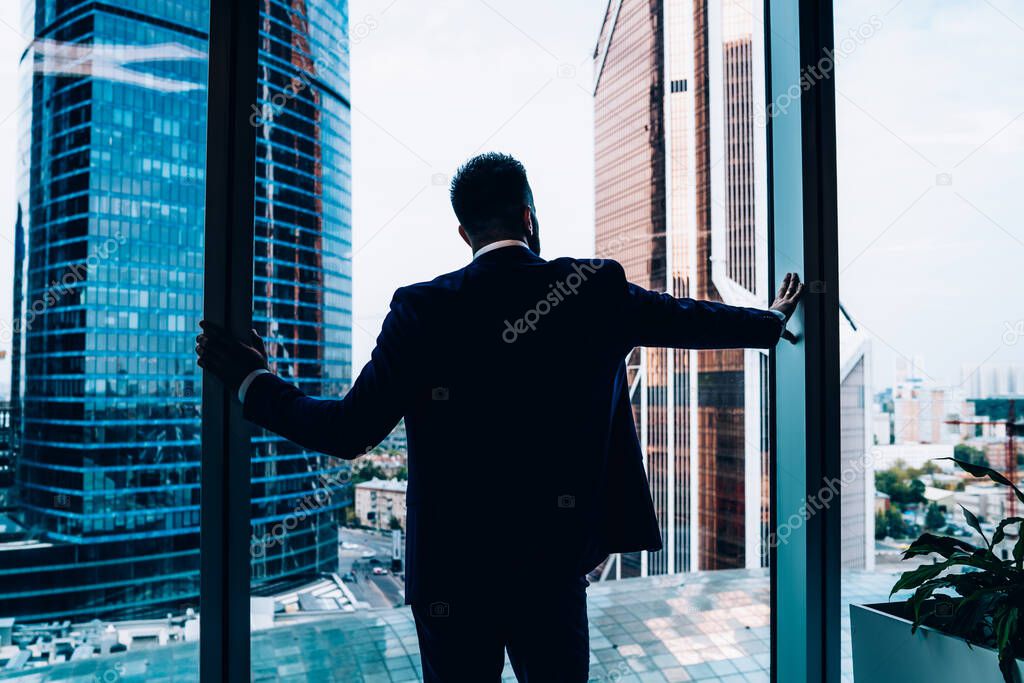 Back view of confident successful businessman in suit leaning on and looking from panoramic window at city center while standing in office