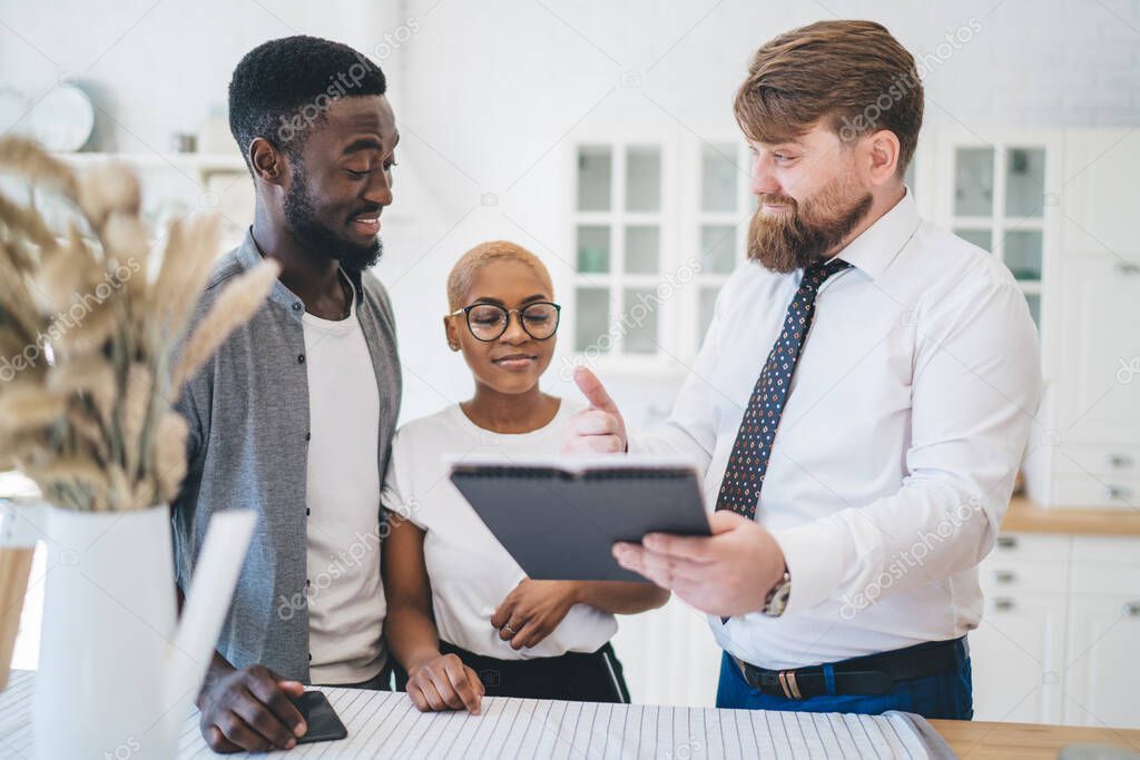 Smiling African American married couple listening to real estate agent offering flat and pointing on mortgage agreement on background of white kitchen 