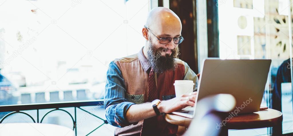 Young cheerful focused spectacled bearded bald man in stylish shirt typing on laptop while sitting in cafe with cup of coffee at daytime