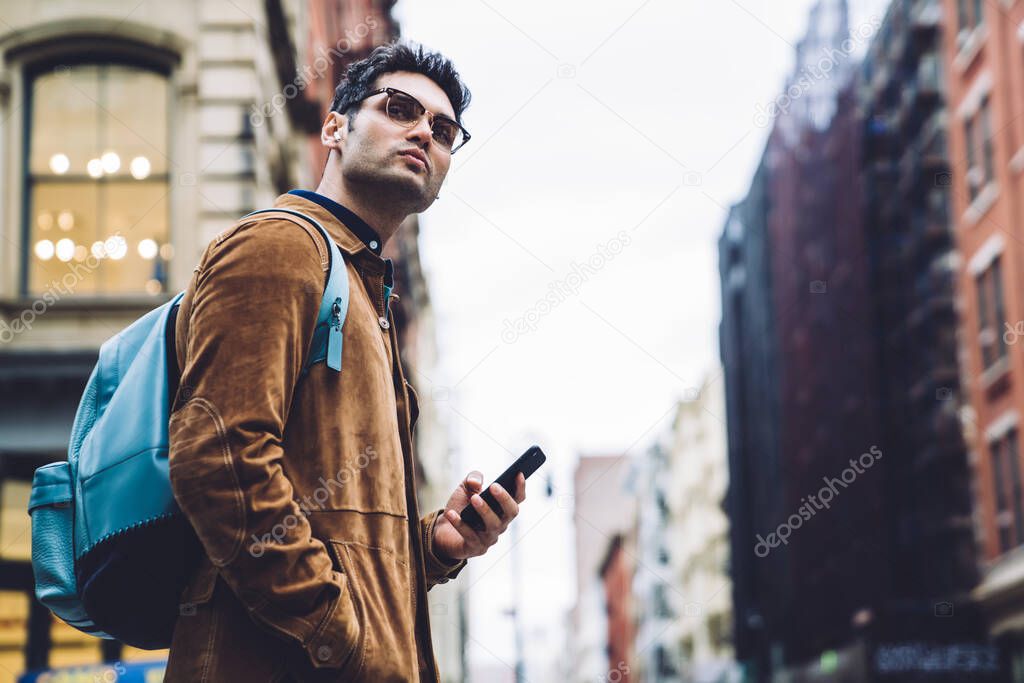 Stylish Hispanic male in eyeglasses wearing brown suede jacket with backpack and earphones standing on crosswalk in New York city and looking away
