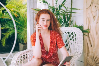 Çekici, dolgun, kırmızı benekli elbiseli, cep telefonuyla bahçe sandalyesinde oturan ve kameraya bakan kızıl saçlı kadın.