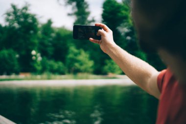 Tanımlanamayan bir adam, yazın parkta gölün bulanık arka planında dururken modern akıllı telefon kullanarak selfie çekiyor.