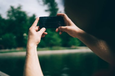Tanımlanamayan adam, güneşli yaz gününde yeşil parkta dinlenirken bulanık göletin fotoğrafını çekmek için modern akıllı telefonu kullanıyor.