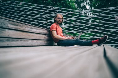 Sıradan kıyafetler ve gözlüklü genç hippi, boş bir tahta tribünde oturuyor ve dizüstü bilgisayarla kameraya bakıyor.