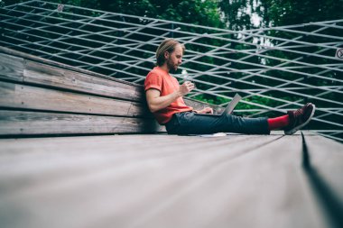 Düşük açılı, rahat, gözlüklü, uzaktan kumandayla çalışan, parktaki ahşap platformda oturan genç bir adam. 