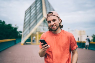 Günlük kıyafetleri ve şapkası olan neşeli erkek akıllı telefonu sosyal ağda gezinmek için kullanıyor. Şehir sokaklarında kameralara bakıyor.