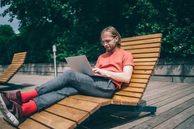 Kırmızı tişörtlü, uzun saçlı, sırıtan kızıl sakallı adam dizüstü bilgisayarlı kırmızı çoraplarla sokakta güneşleniyordu.