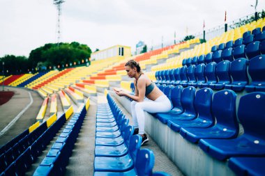 Modern spor kıyafetli, renkli stadyum tribününün sandalyesinde oturan ve gezici gezgin bir kadının yan görüntüsü. 