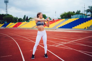 Beyaz taytlı zayıf bir kadın ve gri bluzlu koşan kırmızı pistte duruyor ve kollarını uzatarak stadyuma bakıyor.