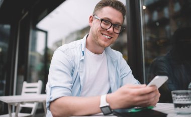 Gözlüklü, mavi tişörtlü, beyaz tişörtlü ve bilekli neşeli entelektüel erkek cep telefonuyla mesajlaşan açık hava terasındaki masada oturuyor.