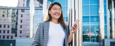 Modern şehir mimarisi arka planında cep telefonuyla ayakta duran başarılı iş kadınının yarım boy portresi, açık havada sohbet etmek için akıllı telefonu kullanan gülümseyen hippi kız.