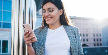 Keyifli esmer ofis çalışanı şık günlük kıyafetler ve moda gözlüklerle açık havada cep telefonuyla mesajlaşırken gülümsüyor.