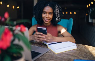 Siyah, neşeli, kıvırcık saçlı, günlük giysili kadın. Kafede sınav hazırlıkları sırasında akıllı telefon kullanarak sosyal medyayı kontrol ediyor.