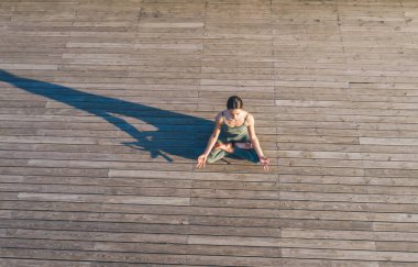 Yoga kıyafetli genç bir kadın ahşap zeminde lotus pozisyonunda oturuyor ve gölgesi düşerek meditasyon yapıyor.